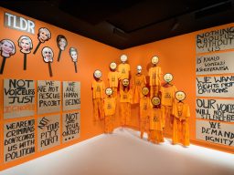 Orangener Raum mit Skulpturen in orangefarbenen Anzügen und kritischen Wandtexten.