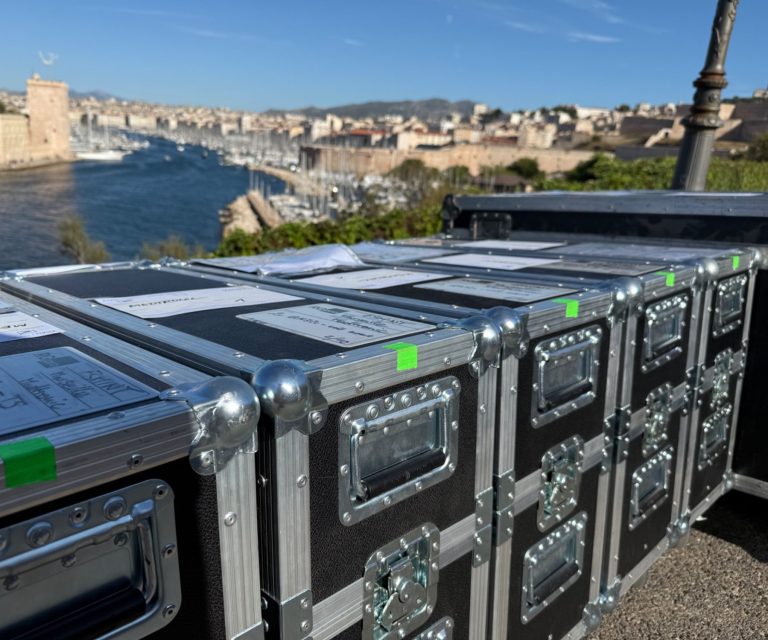 Schwarze Transportkisten mit Aufklebern, Blick auf eine Stadt am Wasser im Hintergrund.