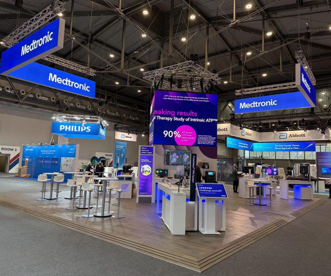 Messestand mit blauen und weißen Displays, Tischen und informierenden Bildschirmen.