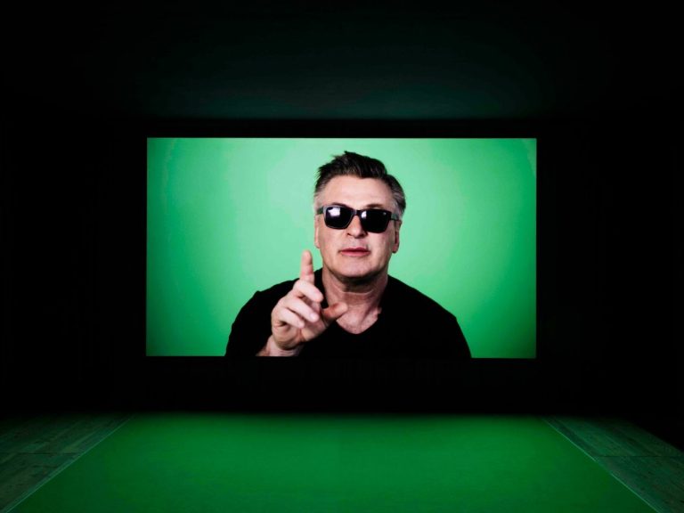 Mann mit Sonnenbrille hält den Zeigefinger hoch vor grünem Hintergrund.